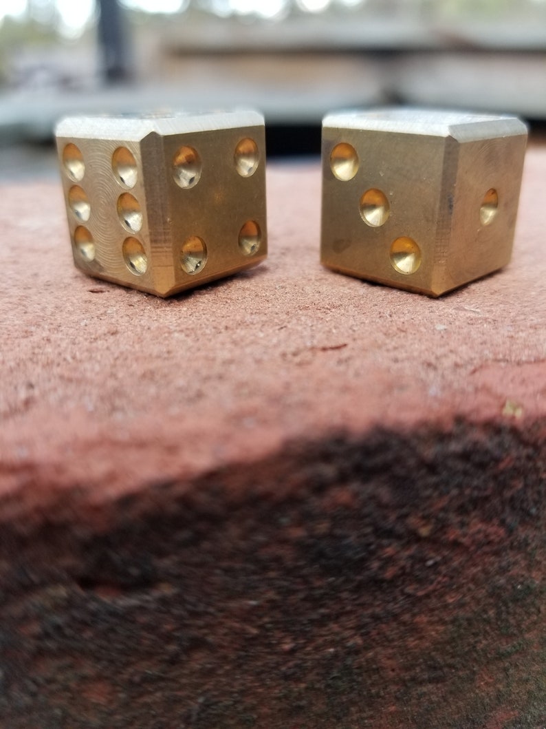 May include: Two gold metal dice with rounded edges. The dice are sitting on a brown surface.