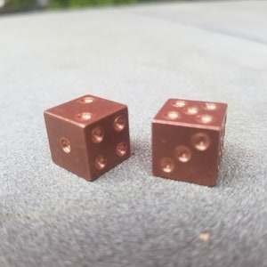 May include: Two copper-coloured dice with rounded edges. The dice are sitting on a grey surface.