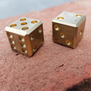 May include: Two gold-colored metal dice with rounded edges. The dice are sitting on a red-brown surface.