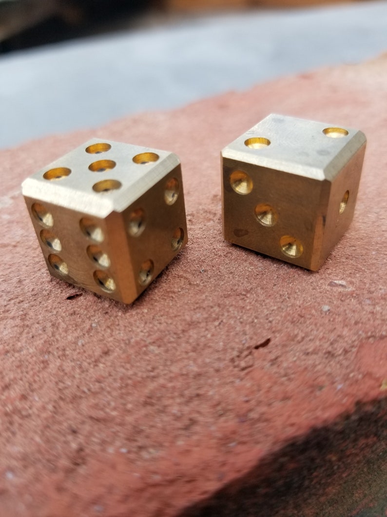 May include: Two gold-colored metal dice with rounded edges. The dice are sitting on a red-brown surface.