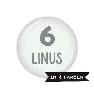Könnte beinhalten: Ein weißer runder Knopf mit einem grauen Tupfenmuster. Die Zahl 6 und der Name Linus sind in Grau auf dem Knopf gedruckt. Der Text "IN 4 FARBEN" ist auf einem schwarzen Band gedruckt.