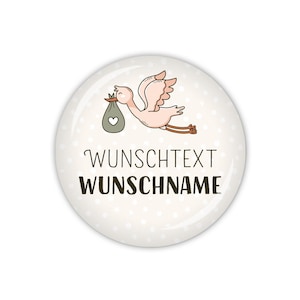 Könnte beinhalten: Ein weißer runder Knopf mit einem rosa Storch, der ein Baby in einem grünen Beutel trägt. Der Knopf hat ein weißes Punkte-Muster und den Text "WUNSCHTEXT WUNSCHNAME" in Schwarz.