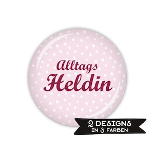 Könnte beinhalten: Ein runder, rosa Button mit weißen Tupfen und den Worten "Alltags Heldinn" in burgunderfarbener Schrift. Ein schwarzes Banner unten zeigt "2 DESIGNS IN 3 FARBEN".