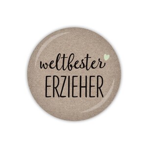 Könnte beinhalten: Ein runder brauner Knopf mit dem Text "weltbester ERZIEHER" und einem kleinen grünen Herz.