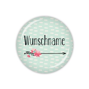 Könnte beinhalten: Ein runder, hellblauer Knopf mit einem weißen und hellblauen Streifenmuster. Der Knopf hat einen schwarzen Pfeil mit rosa Blumen und den Text "Wunschname" in Schwarz.