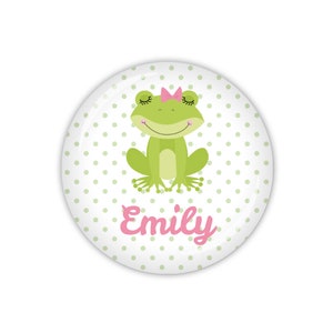 Könnte beinhalten: Ein grüner Frosch mit rosa Wimpern und einer rosa Schleife sitzt auf einem weißen Hintergrund mit grünen Punkten. Der Name "Emily" ist in rosa unter dem Frosch geschrieben.