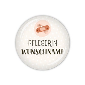 Könnte beinhalten: Weißer runder Knopf mit einem rosa Kreis und einem braunen Pflaster in der Mitte. Der Text "PFLEGERIN WUNSCHNAME" ist in Schwarz auf dem Knopf gedruckt.