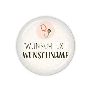 Könnte beinhalten: Ein weißer runder Knopf mit einem rosa Kreis in der Mitte. Der rosa Kreis enthält ein Cartoon-Bild eines Stethoskops. Der Knopf hat weiße Punkte auf weißem Hintergrund. Der Text "WUNSCHTEXT WUNSCHNAME" ist in Schwarz auf dem Knopf gedruckt.