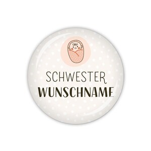 Könnte beinhalten: Weißer Knopf mit rosa Punkten und einem Cartoon-Baby in einer rosa Decke. Der Text "SCHWESTER WUNSCHNAME" ist in Schwarz gedruckt.