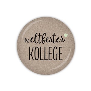 Könnte beinhalten: Ein runder brauner Knopf mit dem Text "weltbester KOLLEGE" und einem kleinen grünen Herzen.