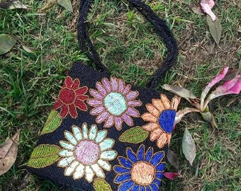 May include: A square handbag featuring a black beaded base decorated with a colorful floral pattern. The flowers are rendered in shades of red, pink, orange, blue, and white with green leaves. It includes two thin black beaded handles.