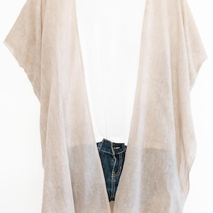 May include: A beige linen kimono-style cover-up draped over a white t-shirt and denim shorts. The cover-up is lightweight and has wide sleeves. The word "FAVORITE" is in a white box with a heart icon.