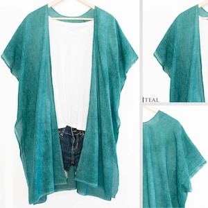 May include: A teal-colored, sheer kimono-style cover-up with short sleeves, draped over a white t-shirt and denim shorts. The cover-up is hanging on a wooden hanger. The word "TEAL" is visible in the image.
