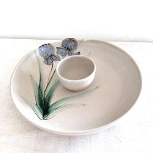May include: A round, off-white ceramic serving dish with an integrated small bowl in the centre. The dish features a hand-painted illustration of two blue iris flowers with long, pointed green leaves extending across the surface of the plate.