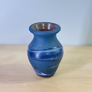 May include: A small ceramic vase with a matte dark blue finish and a wide, rounded rim. The exterior features a swirling, textured white and lighter blue brushstroke pattern that wraps around the body, while the interior reveals a smooth brown clay.