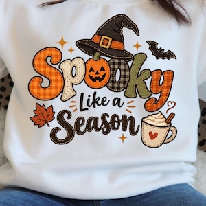 May include: A white sweatshirt featuring a graphic with the text Spooky Like a Season. The design includes a jack-o'-lantern wearing a witch hat, a maple leaf, a bat, and a mug of hot cocoa with a heart, all in orange, brown, and black.