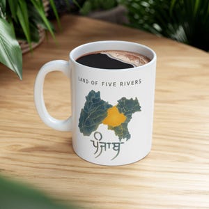 May include: A white ceramic mug filled with dark coffee, featuring a map graphic of the Punjab region with one section highlighted in yellow. The text "LAND OF FIVE RIVERS" is printed above the map, and the word "ਪੰਜਾਬ" is printed below it.