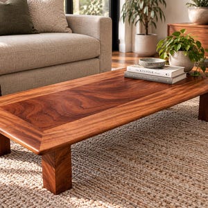 May include: A low-profile rectangular coffee table crafted from rich, reddish-brown wood with a prominent grain pattern. The table features a smooth, flat surface with a wide border and four sturdy, block-style legs, showcasing natural wood tones.