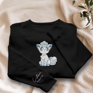 May include: A black long-sleeve sweatshirt featuring a central graphic of a white and light blue cartoon fox-like creature with large eyes. The left sleeve includes the text Custom name embroidered in a simple white cursive script font.