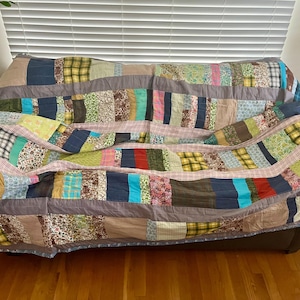 May include: A rectangular patchwork quilt featuring a variety of fabric strips in diverse colors and patterns, including floral, plaid, and solid textures. The quilt is constructed with horizontal grey fabric bands separating the colorful sections.