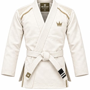 Puede incluir: Un gi de artes marciales color blanco hueso con un logotipo de corona dorada en el pecho izquierdo y los hombros. La chaqueta incluye un cinturón a juego con un pequeño parche de corona y tres rayas negras. La etiqueta interior dice RBI WEARS y A2.