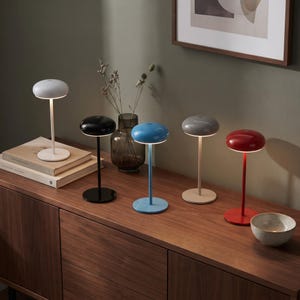 May include: Five portable table lamps with mushroom-shaped shades in white, black, blue, gray, and red. Each lamp features a thin vertical stem and a circular base. The lamps are arranged in a row, emitting a soft light from beneath the shade.