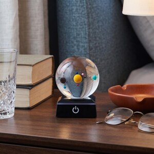 Puede incluir: Una esfera de cristal transparente que contiene un modelo en miniatura del sistema solar con planetas coloridos orbitando alrededor de un sol amarillo central. La esfera descansa sobre una base cuadrada negra con un icono de encendido.