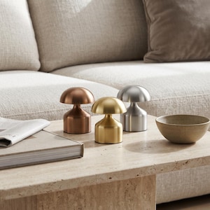 Könnte beinhalten: Drei kleine pilzförmige Tischlampen mit glatten Metalloberflächen in Kupfer, Gold und Silber. Jede Lampe verfügt über einen abgerundeten Kuppelschirm und einen zylindrischen Sockel und steht zusammen auf einer flachen Oberfläche neben einem Buch.