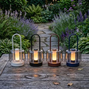 May include: Four portable table lamps with arched handles and cylindrical glowing light cores. Each lamp features a clear glass dome shade and a solid circular base. The lamps are finished in white, black, brown, and dark blue, arranged in a row.