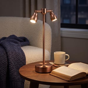 May include: A copper-toned metal desk lamp featuring a central vertical stand and two adjustable, downward-facing spotlight heads. The lamp has a circular base with a small power switch and emits a warm, focused light from both light fixtures.