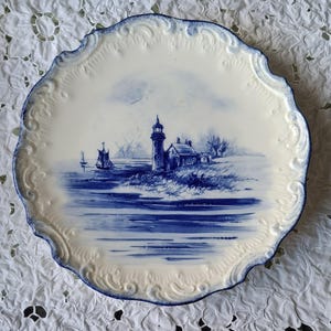 Może przedstawiać: Okrągły talerz ceramiczny z falistym brzegiem, ozdobiony niebiesko-białym wzorem. W centrum znajduje się latarnia morska na skalistym brzegu oraz żaglówka na wodzie. Brzeg talerza posiada wypukły wzór z niebieskimi akcentami kolorystycznymi.