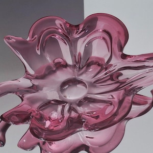 Puede incluir: Un cuenco de cristal magenta translúcido con forma de flor abierta, con múltiples pétalos curvos y ondulados que irradian desde una base circular central. El cristal presenta líneas fluidas y un acabado brillante que refleja la luz.