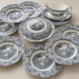 Op de afbeelding: Een set keramisch serviesgoed met een donkerblauw en wit transferpatroon van een kasteellandschap. De set bevat diverse ronde borden, een serveerschaal met deksel en decoratieve handgreep, en een sauskom op een bijpassende schotel.
