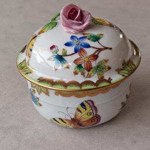 Könnte beinhalten: Eine weiße Porzellanschale mit Deckel, verziert mit handgemalten bunten Schmetterlingen und Blumenmotiven. Der Deckel ist mit einer skulptierten rosa Rose gekrönt und mit Goldverzierungen an den Rändern versehen.