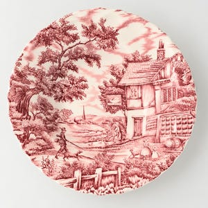 Può includere: Un piatto rotondo in ceramica con un motivo a trasferimento rosso e bianco. Il design raffigura una scena rurale con un edificio chiamato The Swan, alberi, una recinzione e figure che camminano con cani in un paesaggio con bordo smerlato.