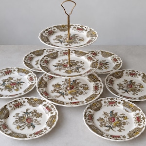 Può includere: Un'alzata a tre piani con manico in metallo color ottone e sette piatti in ceramica abbinati. Ogni piatto presenta una base bianca con bordo smerlato e un motivo floreale centrale in rosso, giallo e blu, accentuato da volute marroni.