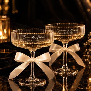 May include: Two clear glass coupe glasses with a vertical ribbed texture, each featuring a white satin ribbon tied around the stem. The glasses are engraved with the text Emma & James and the date 10.05.2026 in a white script and sans-serif font.