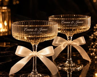 May include: Two clear glass coupe glasses with a vertical ribbed texture, each featuring a white satin ribbon tied around the stem. The glasses are engraved with the text Emma & James and the date 10.05.2026 in a white script and sans-serif font.