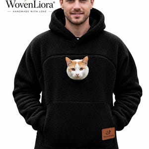 Puede incluir: Una sudadera con capucha de forro polar negro que presenta un gran bolsillo frontal tipo canguro con un gráfico realista de una cara de gato blanco y naranja centrado en el pecho. La prenda incluye capucha, mangas largas y una pequeña etiqueta marrón.