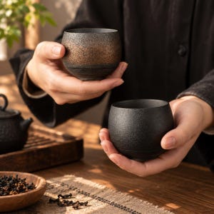 May include: Two small, round ceramic tea cups with a textured finish. One cup features a dark charcoal base with a metallic bronze band around the middle, while the other cup is a solid matte black with a rough, granular surface texture.