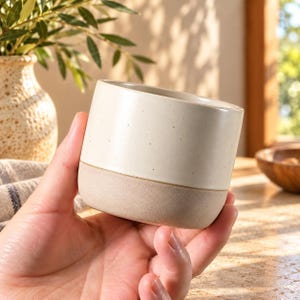 Puede incluir: Una pequeña taza cilíndrica de cerámica con un diseño de dos tonos. La parte superior tiene un esmalte blanco roto liso con sutiles motas oscuras, mientras que la base inferior presenta una textura beige mate sin esmaltar.