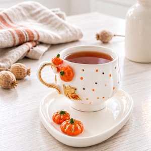 Op de afbeelding: Een witte keramieken theekop en schotel set met kleine oranje pompoen- en goudkleurige bladaccenten. De kop heeft een goudkleurig handvat en is versierd met kleine oranje en bruine stippen. Twee kleine oranje pompoenen liggen op de schotel.
