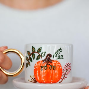 Könnte beinhalten: Eine weiße Keramiktasse mit passender Untertasse, verziert mit einem handgemalten orangefarbenen Kürbis mit braunem Stiel, grünen Ranken und roten Beeren. Die Tasse verfügt über einen markanten, kreisförmigen goldfarbenen Henkel und eine glatte Oberfläche.