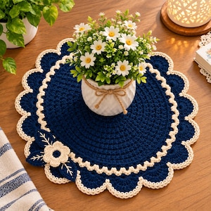 Könnte beinhalten: Ein rundes, dunkelblaues Häkeldeckchen mit einem cremefarbenen, gewellten Rand und einer dekorativen cremefarbenen Blumenapplikation mit Blättern. Das Deckchen liegt unter einem weißen Keramiktopf mit weißen Gänseblümchen mit gelber Mitte.