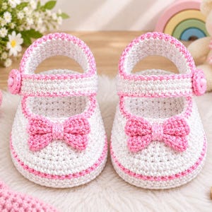 Könnte beinhalten: Ein Paar gehäkelte Babyschuhe in Weiß mit rosa Rand, einer rosa Schleife auf der Vorderseite und einem Riemen mit rosa Knopfverschluss. Die Schuhe sind in einem strukturierten Maschenmuster gefertigt und haben eine abgerundete Zehenpartie.