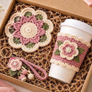 Könnte beinhalten: Eine Geschenkbox mit einem runden Häkeluntersetzer mit rosa, grünen und cremefarbenen Blumenmustern, einem weißen Kaffeebecher mit passender rosa-cremefarbener Häkelmanschette mit Blume und einem Häkel-Schlüsselanhänger mit Blume und Quaste.