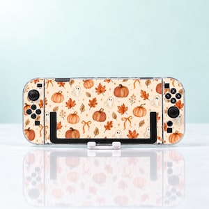 May include: A handheld gaming console featuring a decorative skin with a repeating pattern of orange pumpkins, brown autumn leaves, small white ghosts, and tan ribbons on a cream background, held in a black plastic stand.