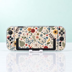 May include: A handheld gaming console featuring a cream-colored skin with a repeating pattern of colorful butterflies, red hearts, yellow crescent moons, sunflowers, various green ferns, and small blue flowers, held in a black plastic stand.