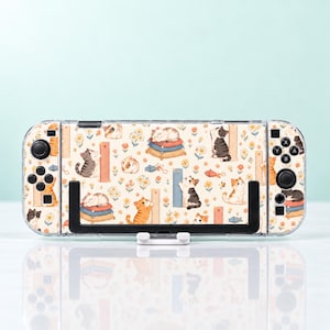 May include: A handheld gaming console featuring a cream-coloured skin with a repeating pattern of various cats, small yellow flowers, scratching posts, and toy mice. The device includes black buttons, joysticks, and a black kickstand on the back.