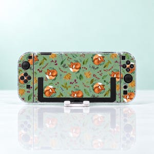 Könnte beinhalten: Eine tragbare Spielkonsole mit einer salbeigrünen Hülle, die mit einem sich wiederholenden Muster aus schlafenden orangefarbenen Füchsen, Herbstblättern und kleinen roten Beeren im Aquarellstil bedruckt ist. Das Gerät hat schwarze Tasten, Joysticks und einen Ständer.
